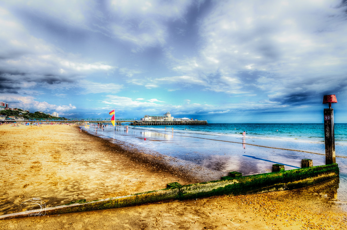Sandy beach with people near the water, a pier extending into the sea under a partly cloudy sky.