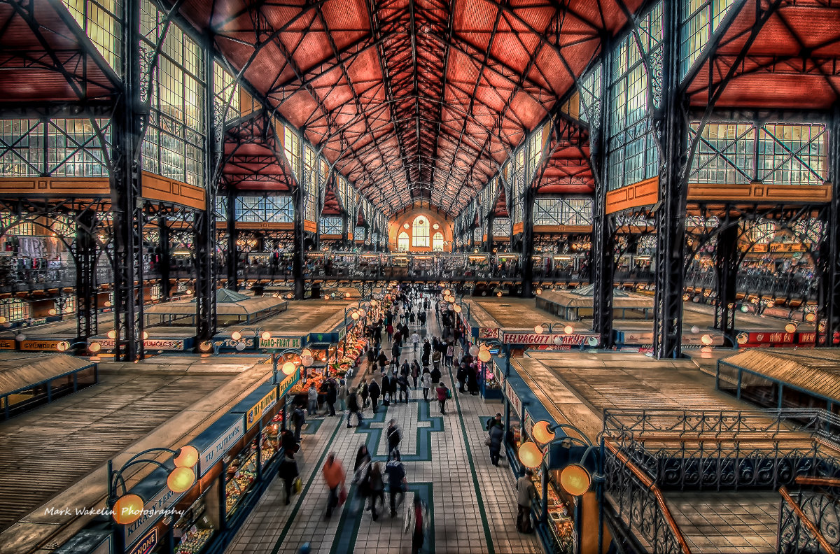 Interior view of a large indoor market hall with vaulted ceiling, tall windows, and rows of stalls with shoppers walking along the tiled central aisle.