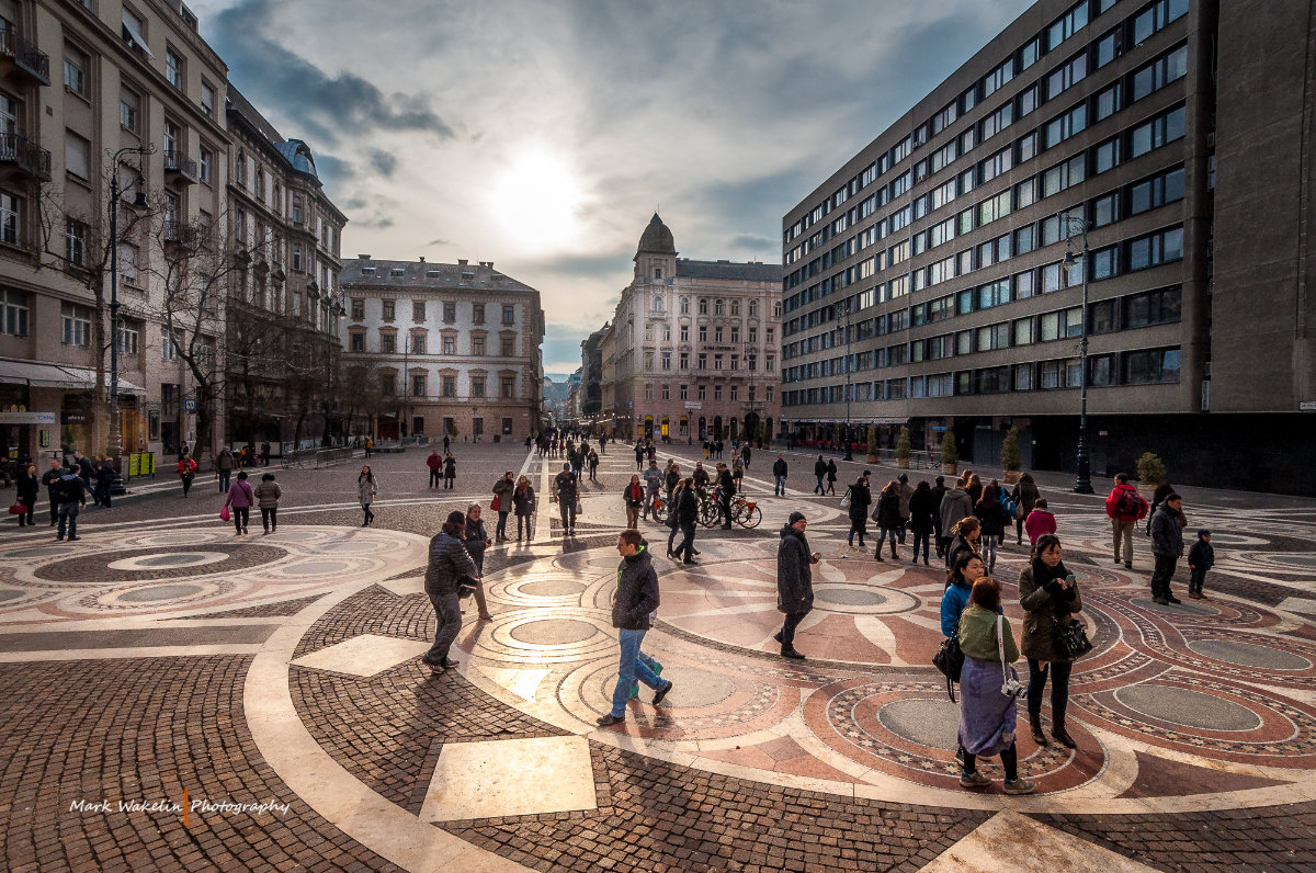 People walking and standing on a large patterned plaza surrounded by historic and modern buildings under a cloudy sky with the sun shining through.