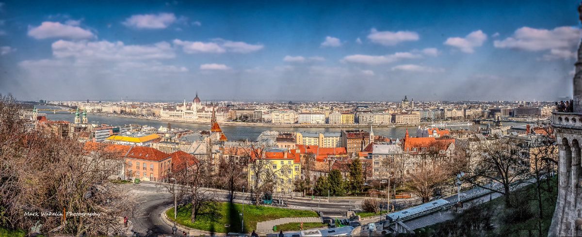 Panoramic view of Budapest featuring the Danube River, the Hungarian Parliament building, and historical cityscape under a partly cloudy blue sky.