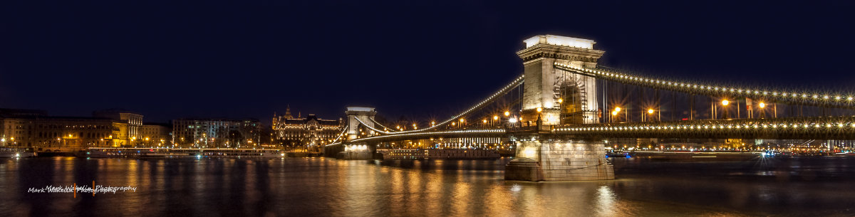 Night view of the illuminated Széchenyi Chain Bridge over the Danube River in Budapest with city lights reflecting on the water.
