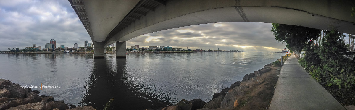 View beneath a concrete bridge over calm water with a city skyline and cloudy sky in the distance, rocks and a walkway with trees on the right.