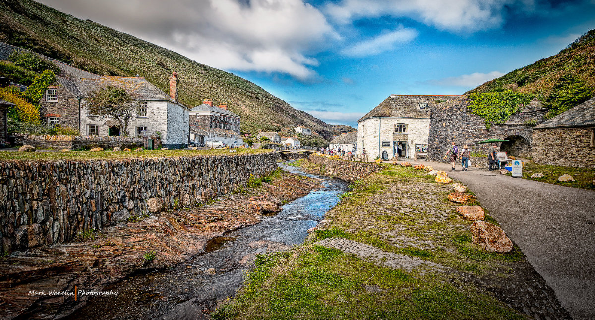 Stone cottages along a narrow stream in a green valley under a partly cloudy sky, with people walking on the path beside.