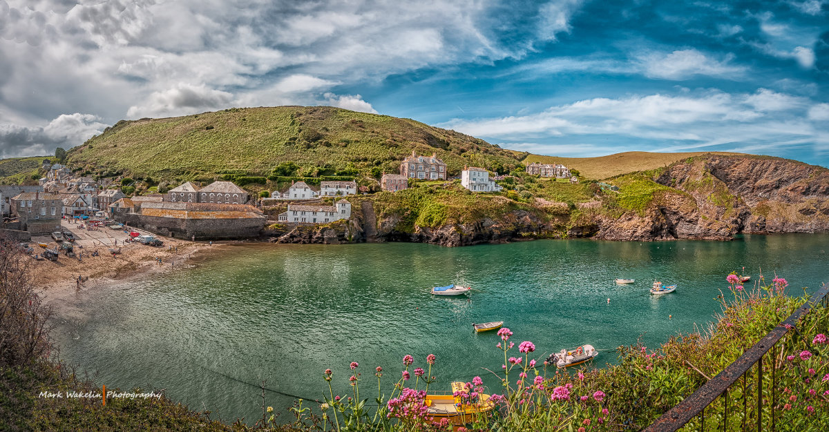 Small coastal village with stone houses beside a calm bay dotted with boats and surrounded by green hills under a partly cloudy sky.