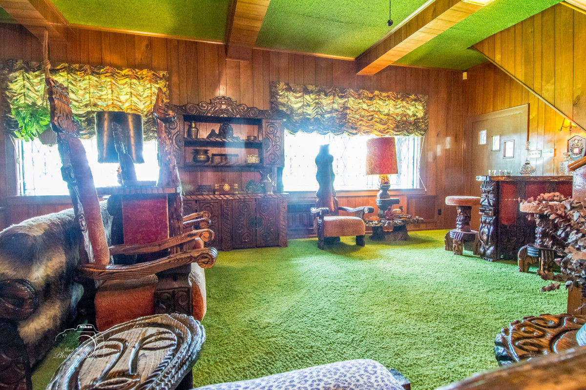 Vintage living room with green carpet, wooden panel walls, carved wooden furniture, and patterned curtains.