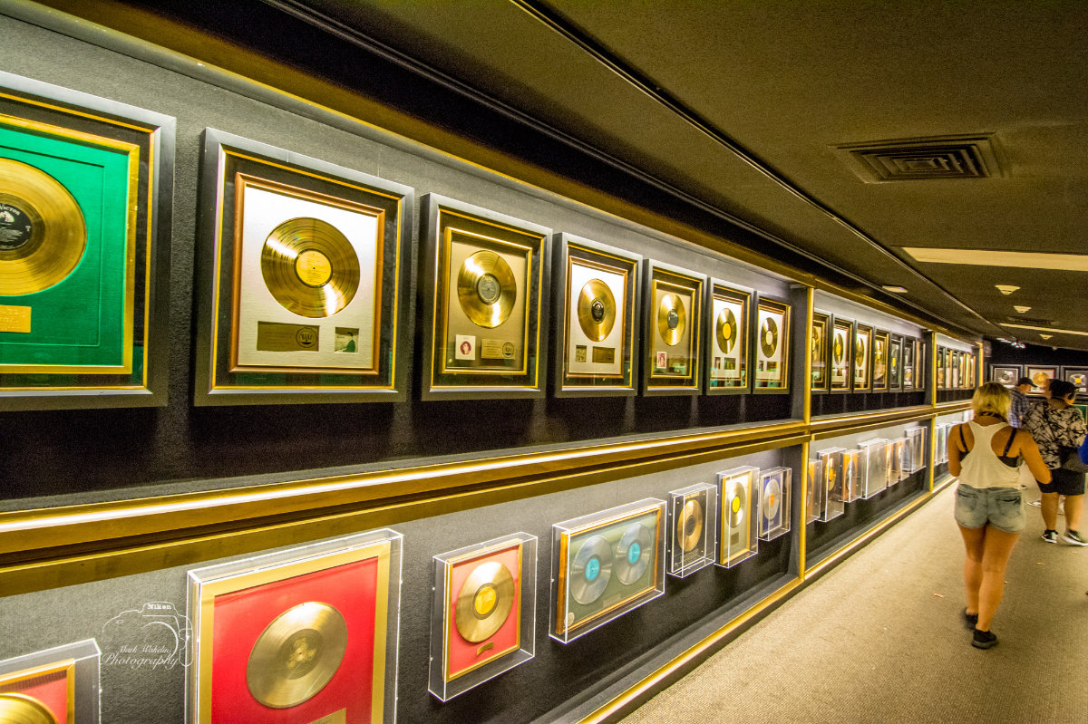 Hallway lined with framed gold and platinum records displayed on dark walls with people walking along.