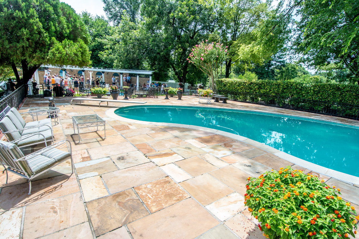 Outdoor swimming pool surrounded by stone tiles, patio furniture, flowering plants, and a crowd of people near a brick building in a garden setting.