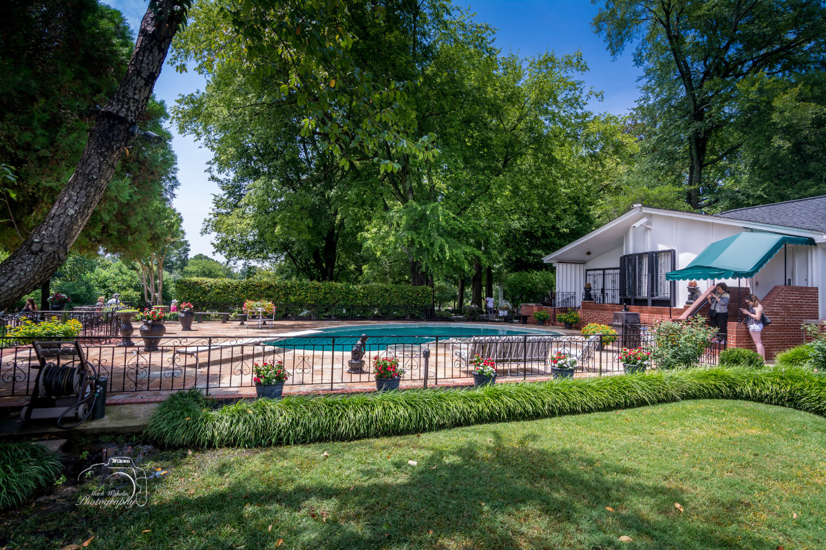 Outdoor pool area surrounded by lush green trees, potted flowers, and a white building with a green awning, featuring people near the entrance.