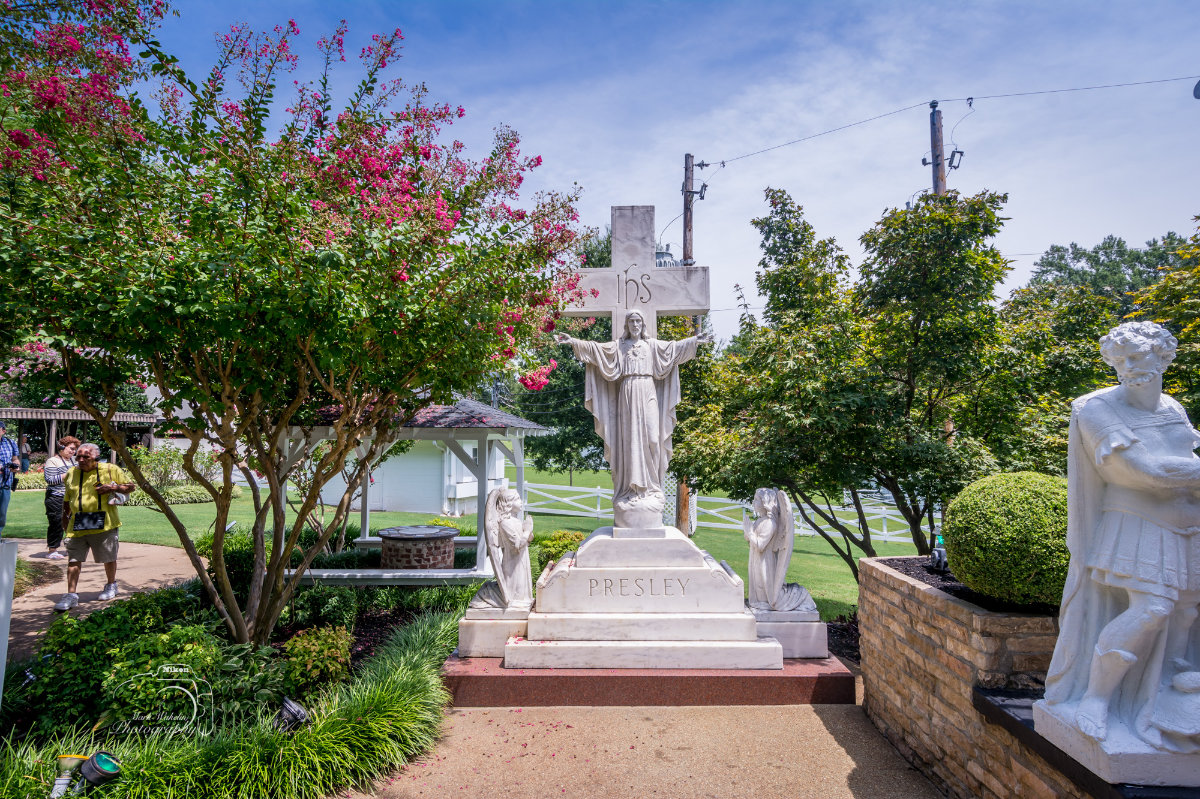 White marble statue of Jesus with outstretched arms in front of a cross inscribed with ‘IHS,’ flanked by angel statues, with Presley engraved below, surrounded by greenery and flowering trees.