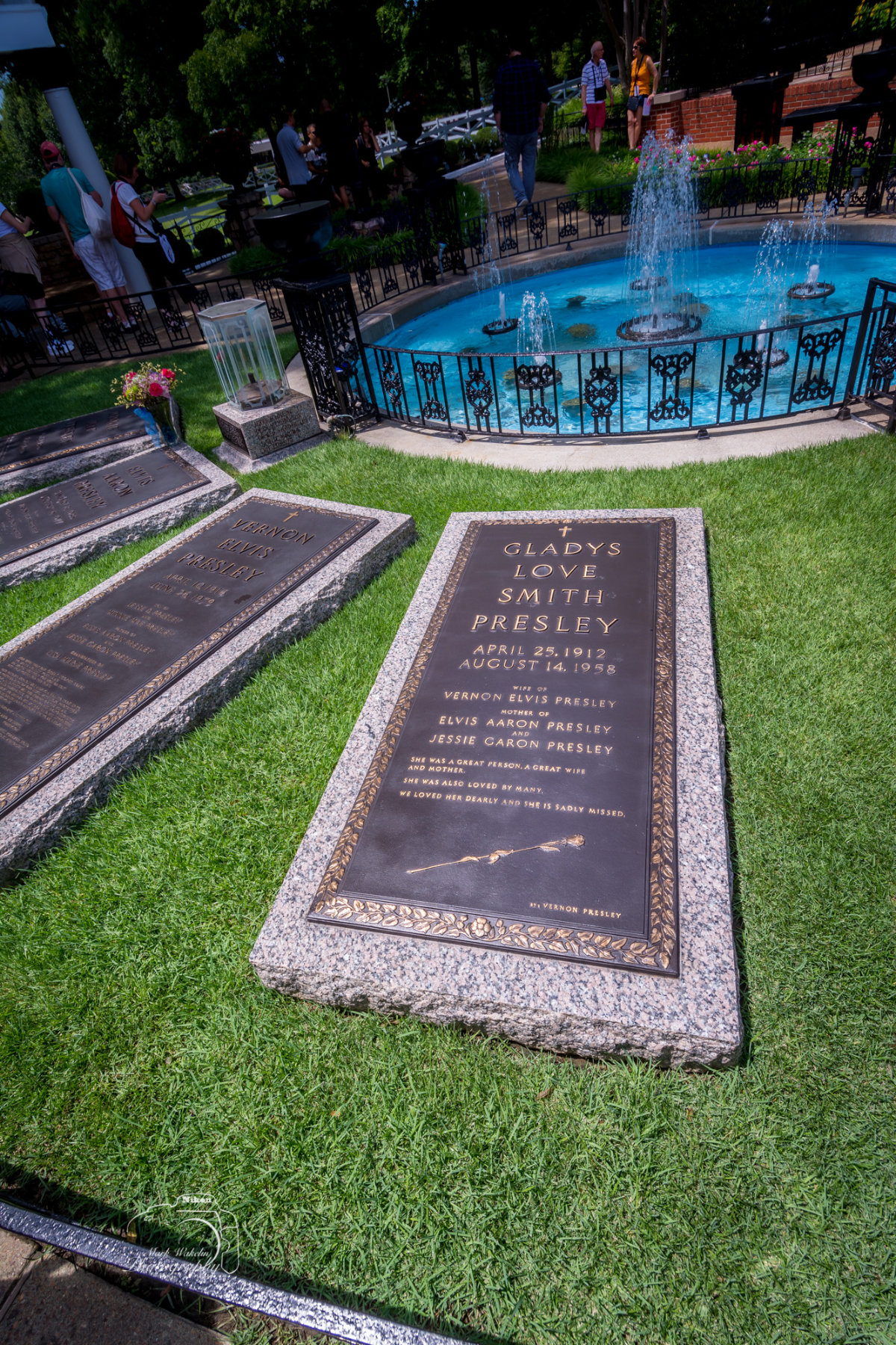 Gravestones of Vernon Elvis Presley and Gladys Love Smith Presley on green grass near a blue fountain with water jets, with visitors in the background.