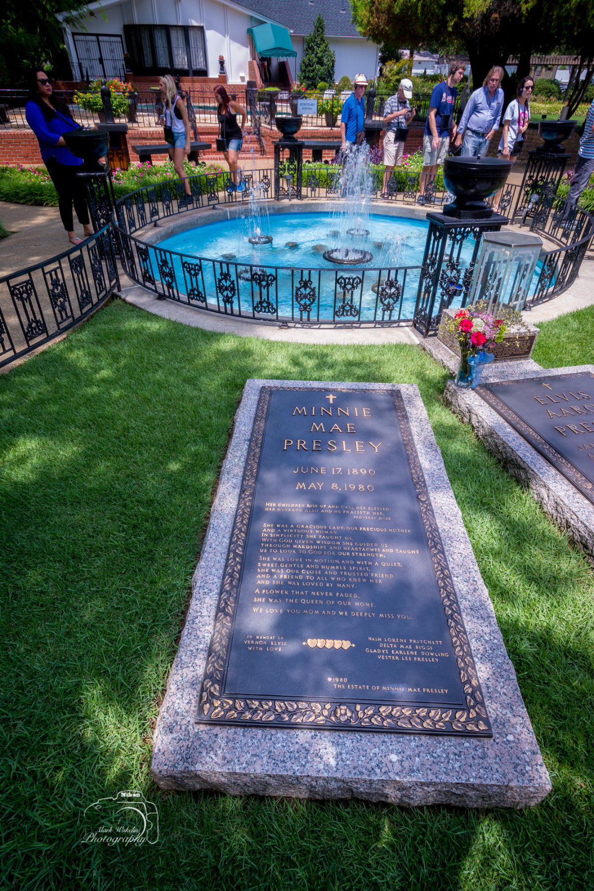 Grave of Minnie Mae Presley with inscription and nearby Elvis Presley grave at Graceland, surrounded by green grass and an ornamental fountain with visitors in the background.