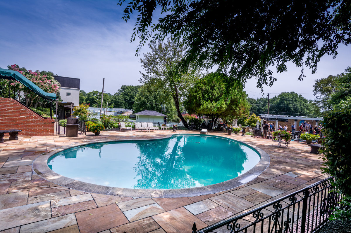 Clear blue swimming pool surrounded by a tiled patio, trees, and outdoor seating on a sunny day.