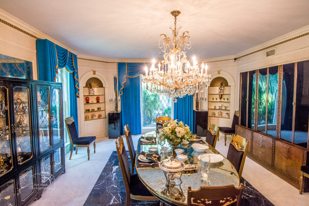 Elegant dining room with a glass table set for six, blue curtains, crystal chandelier, and display cabinets.