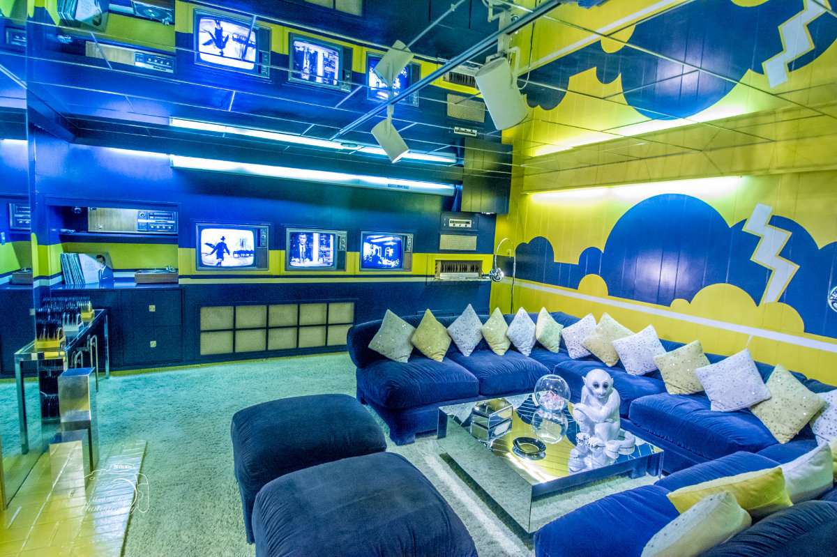 Retro living room with blue and yellow walls, multiple vintage TVs, a large blue sectional sofa with numerous cushions, and a mirrored coffee table with decorative items.