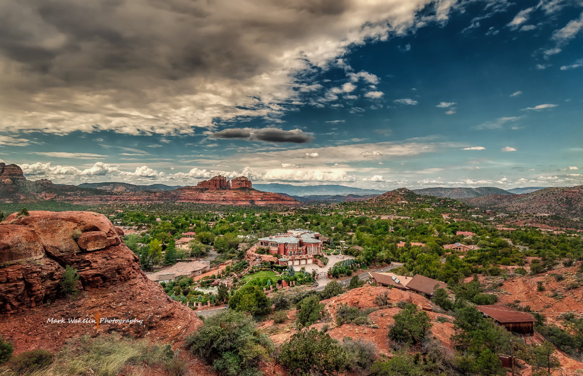 Panoramic view of a red-rock desert landscape with scattered houses and a large mansion under a dramatic cloudy sky.