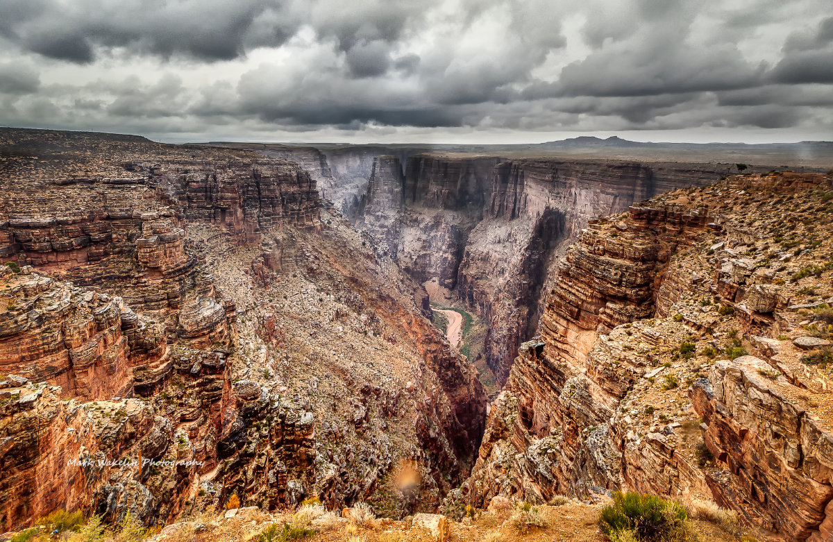 Vast rocky canyon under a cloudy sky with a river flowing at the bottom.