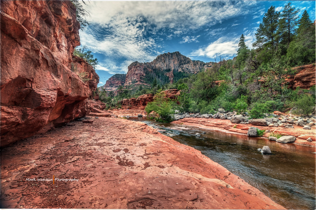 Red rock formations and clear river flowing through a green forested canyon under a partly cloudy blue sky.