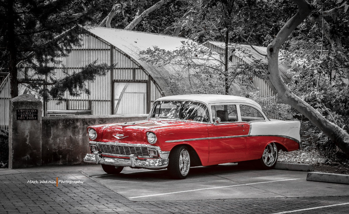 Red and white classic Chevrolet car parked next to a wall with a sign and surrounded by trees.