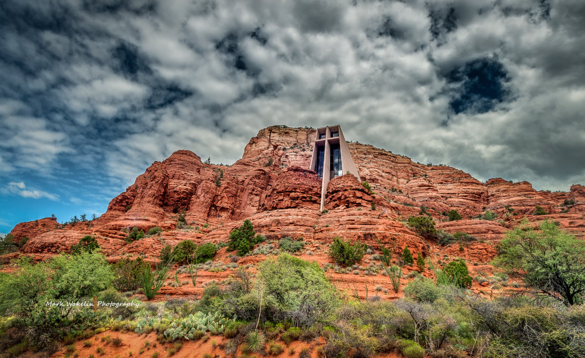 Red rock formations with green vegetation and a modern chapel built into the cliff under a dramatic cloudy sky.