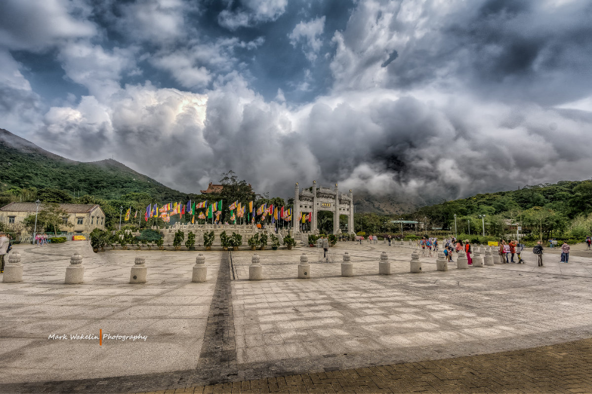 Wide stone-paved plaza with a traditional archway and colorful flags, surrounded by green hills under a dramatic cloudy sky.