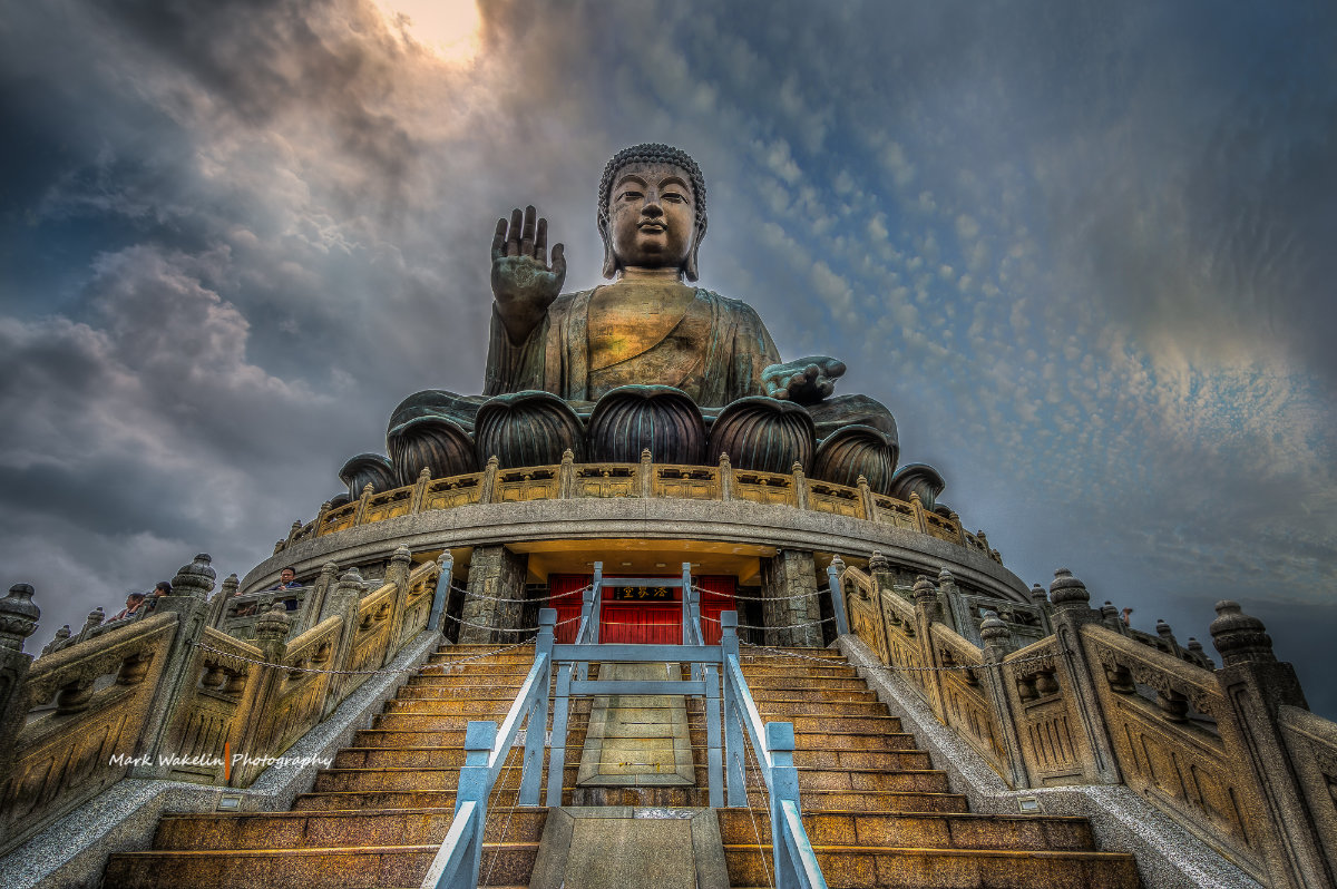 Large bronze Tian Tan Buddha statue atop stone stairs with dramatic cloudy sky background.
