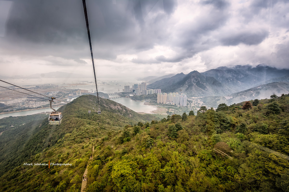 Cable cars travelling over a green mountainous landscape with a city, water and cloudy sky in the background.