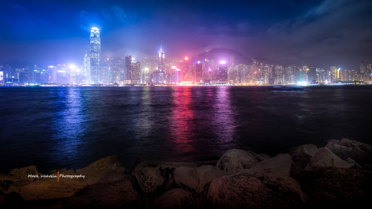 Night view of a brightly lit city skyline reflecting colourful lights on the water with rocks in the foreground.