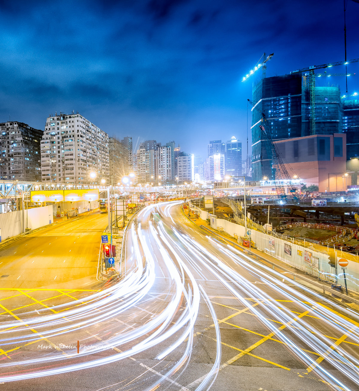 Long exposure shot of light trails from vehicles on a busy city road at night with high-rise buildings and construction cranes in the background.
