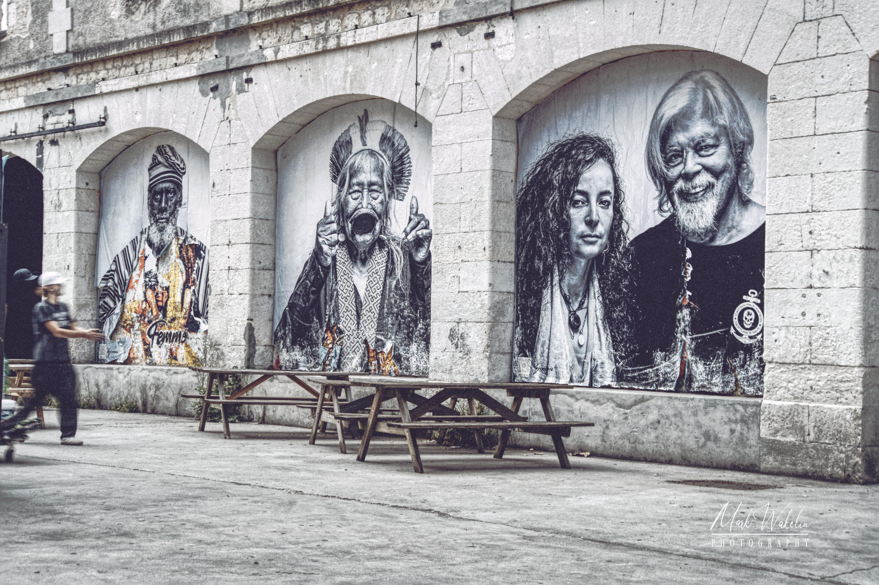 Outdoor wall murals featuring black-and-white portraits of diverse people, with picnic tables in front and a person walking by.