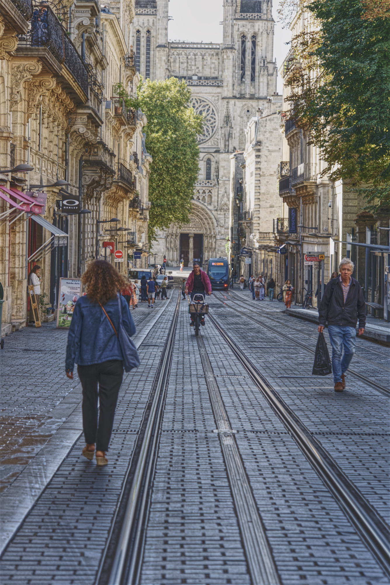 Pedestrians and a cyclist on a cobblestone street with tram tracks, lined with historic buildings and a large cathedral in the background.
