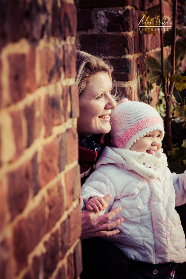 Smiling woman crouched behind a brick wall holding a baby wearing a pink and white knitted hat and white jacket.