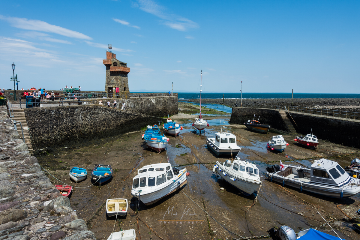 Small boats resting on wet ground in a dry harbour with stone walls and a small stone tower under a blue sky.