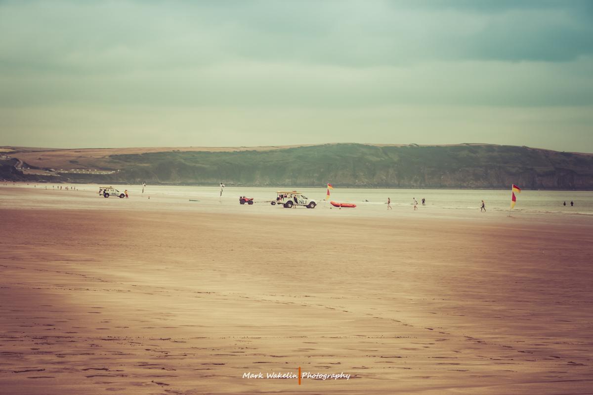Wide sandy beach with two RNLI lifeguard vehicles, rescue boards, red and yellow safety flags, and a few people near the shoreline under an overcast sky.