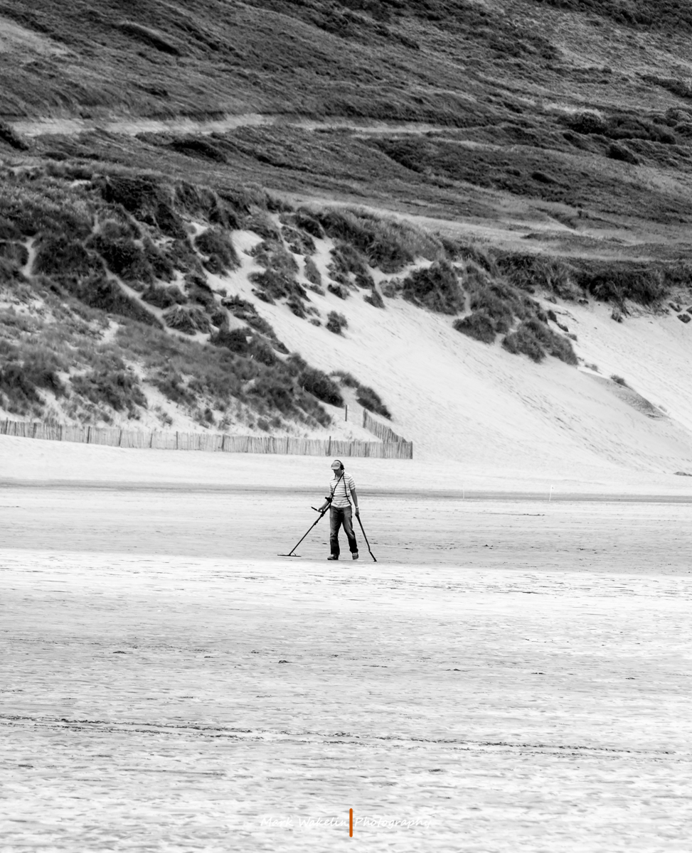 Person using a metal detector on a vast sandy beach with grassy dunes in the background.