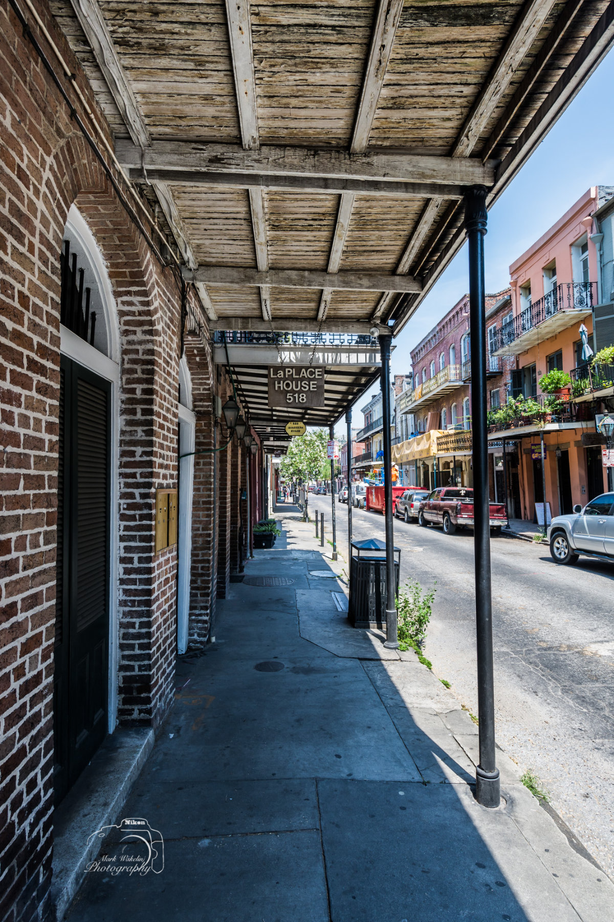 Covered sidewalk with brick building on the left and street with parked cars on the right under a clear blue sky in a historic urban area.
