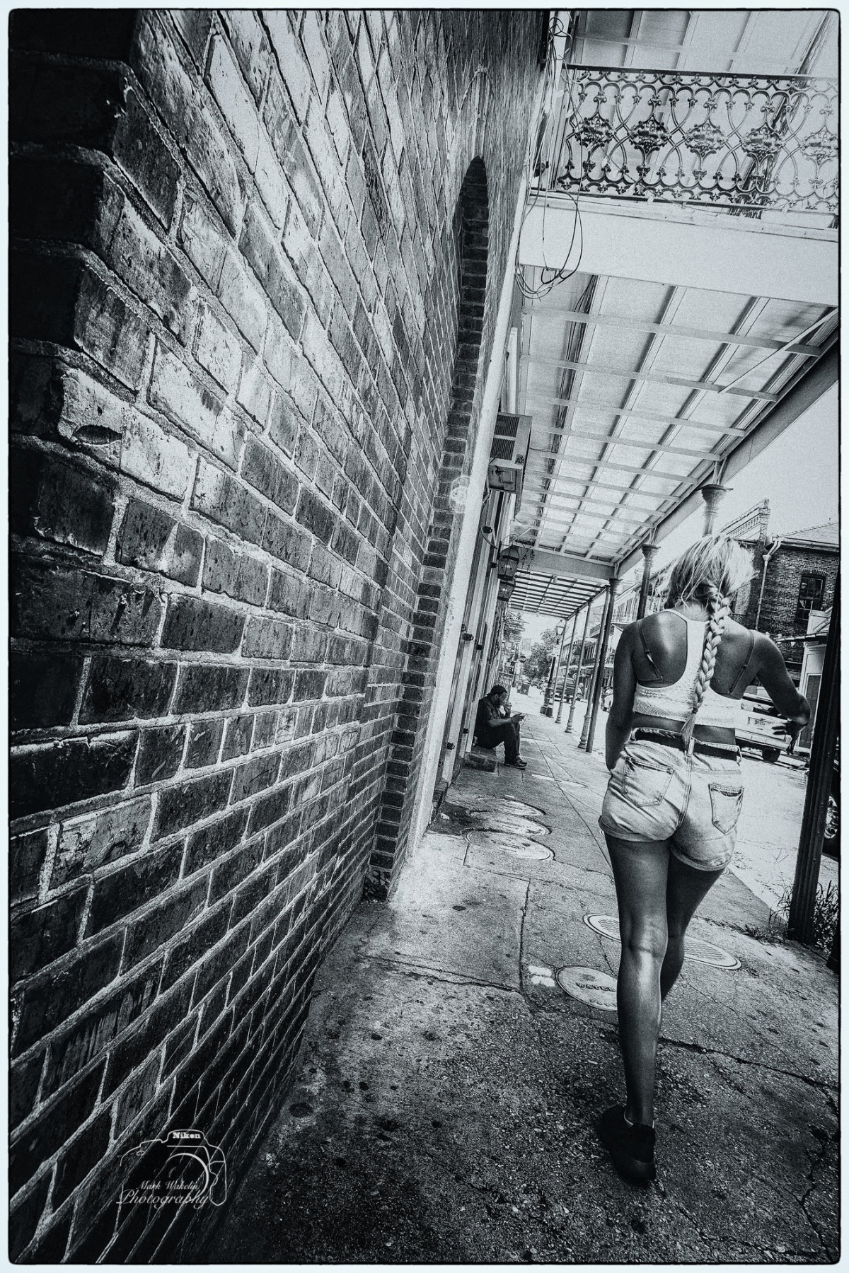 Black and white photo of a woman with a long braid walking along a sidewalk beside a brick wall and a man sitting in the background.