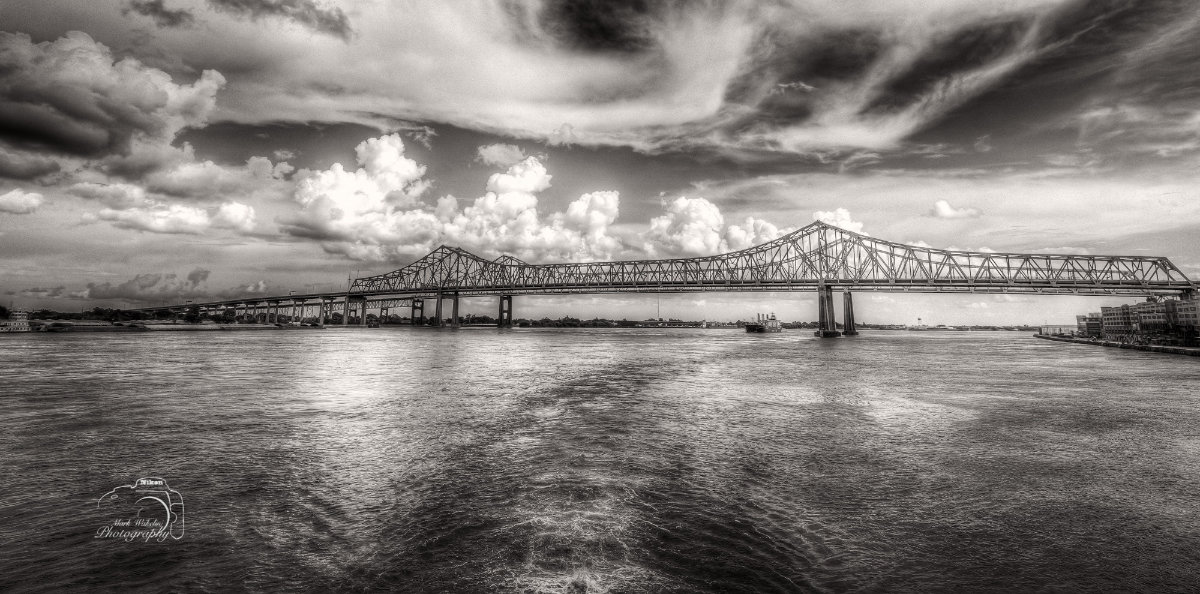 Black and white photo of a large steel truss bridge spanning a wide river under a dramatic cloudy sky.