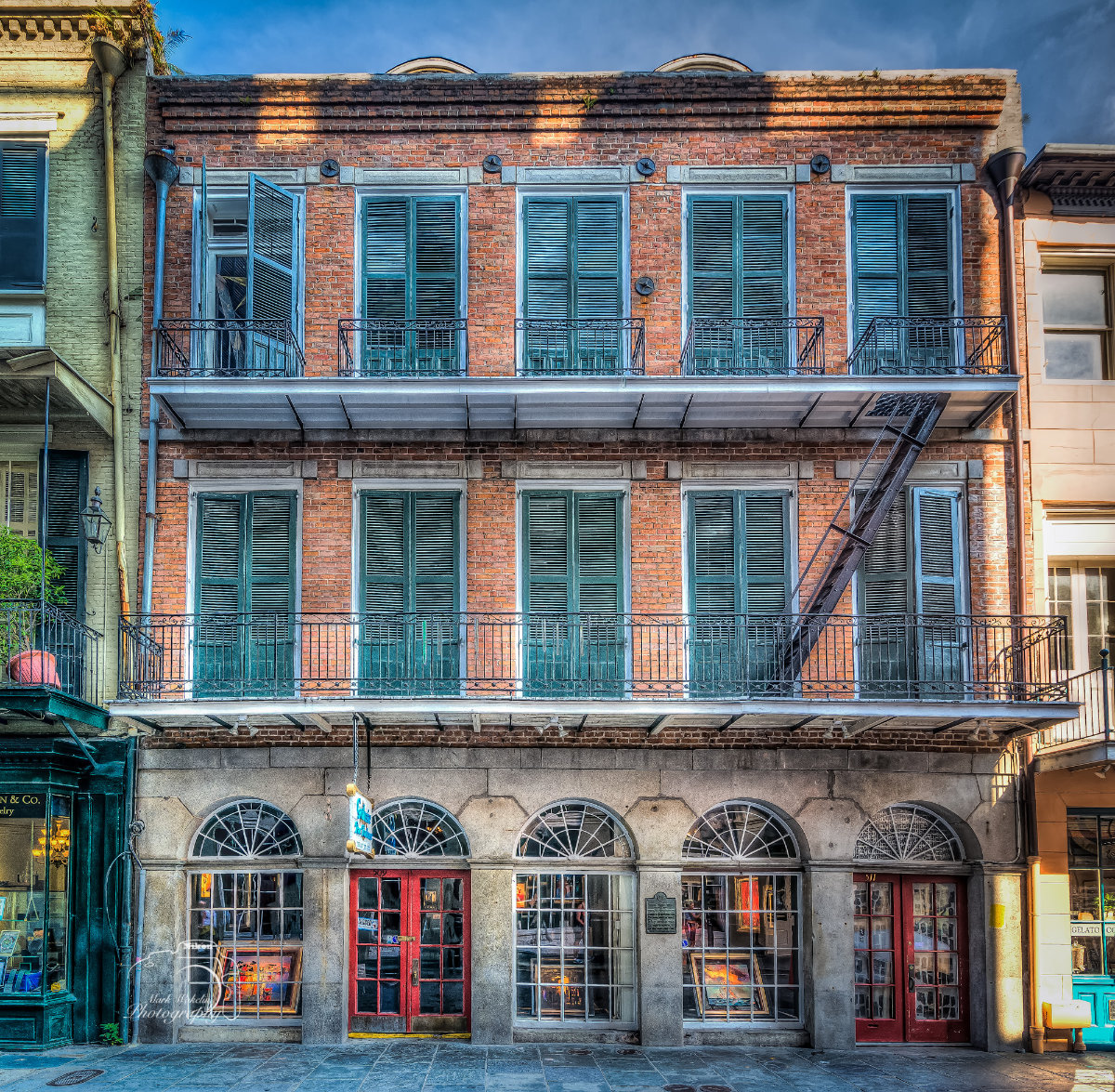 Three-story brick building with blue shutters, balconies, red double doors, and arched windows on the ground floor.