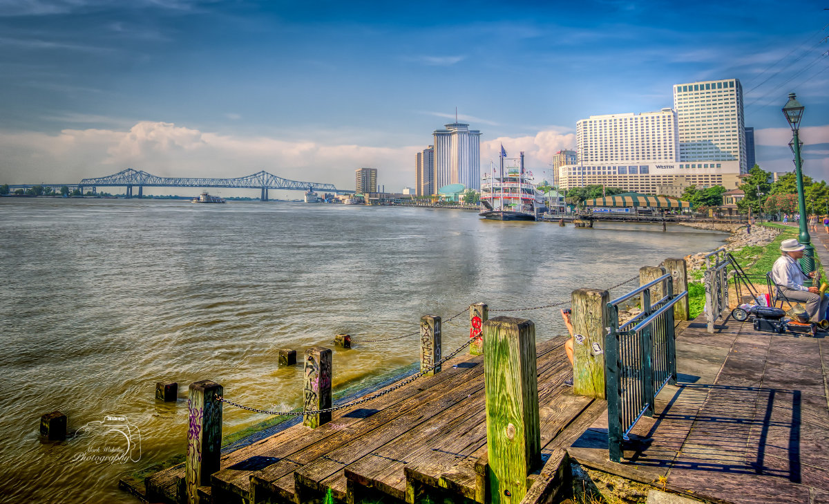 Riverside wooden pier with a seated man playing saxophone, paddle steamboat docked, and city skyline with bridge in the background under a blue sky.