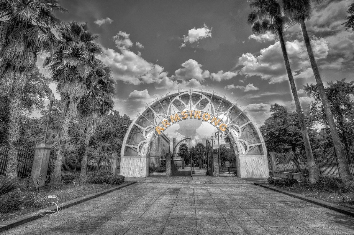 Wide path leading to a decorative archway with the illuminated word 'ARMSTRONG' surrounded by palm trees under a cloudy sky.