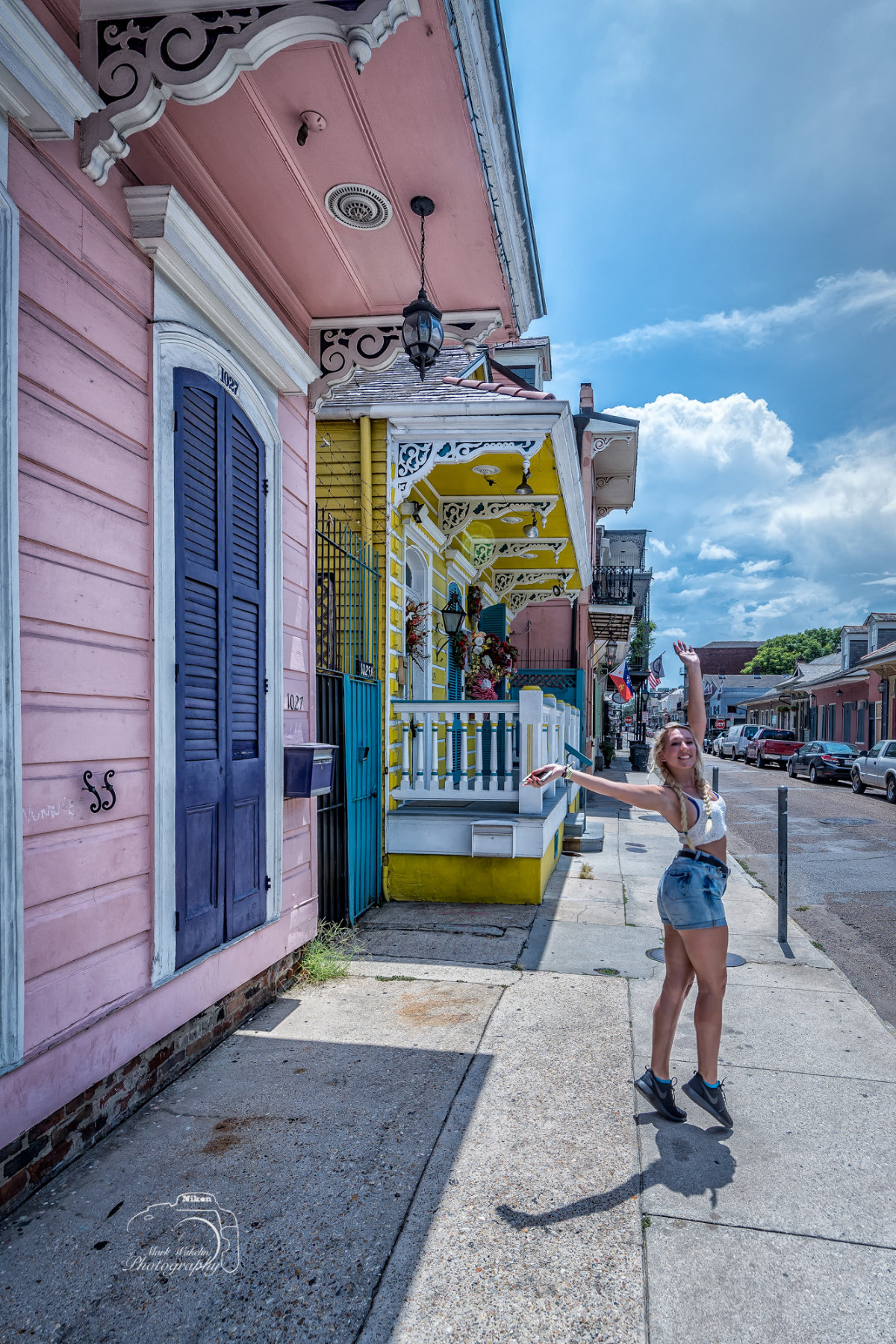 Woman in denim shorts and white crop top jumping joyfully on a sunny sidewalk with colourful New Orleans-style houses in the background.