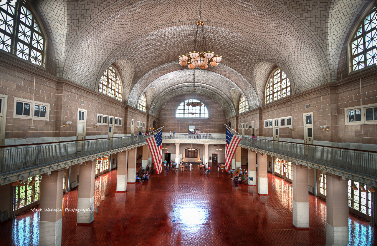 Interior of a large hall with arched tiled ceiling, polished red floor, American flags, and visitors seated along the walls.