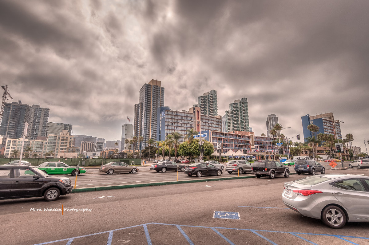 City street with multiple cars and tall buildings under a cloudy sky in an urban area.
