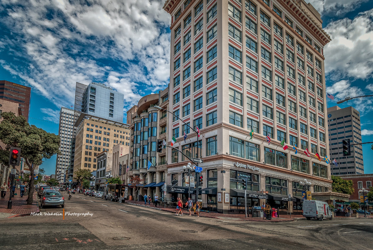 City street corner with a tall building displaying multiple international flags and people walking on the sidewalks under a partly cloudy sky.