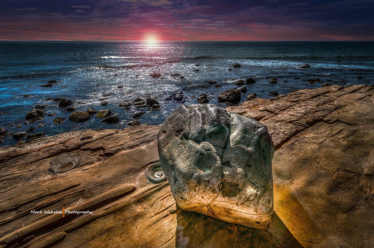 Rocky coastline with a large weathered rock in the foreground and the sun setting over the ocean horizon.