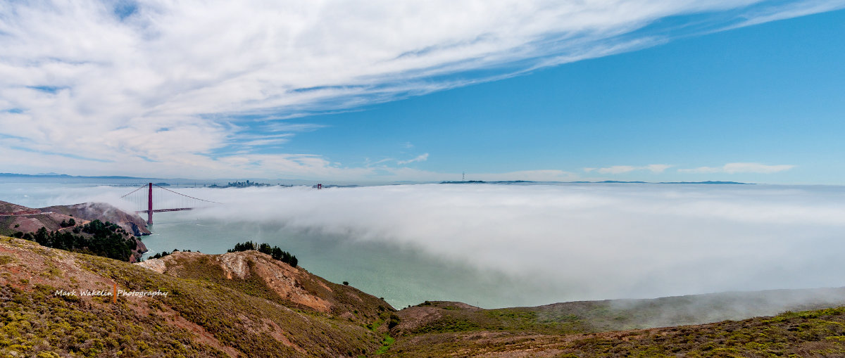 Golden Gate Bridge rising above a dense layer of fog with hills in the foreground and a partly cloudy blue sky.