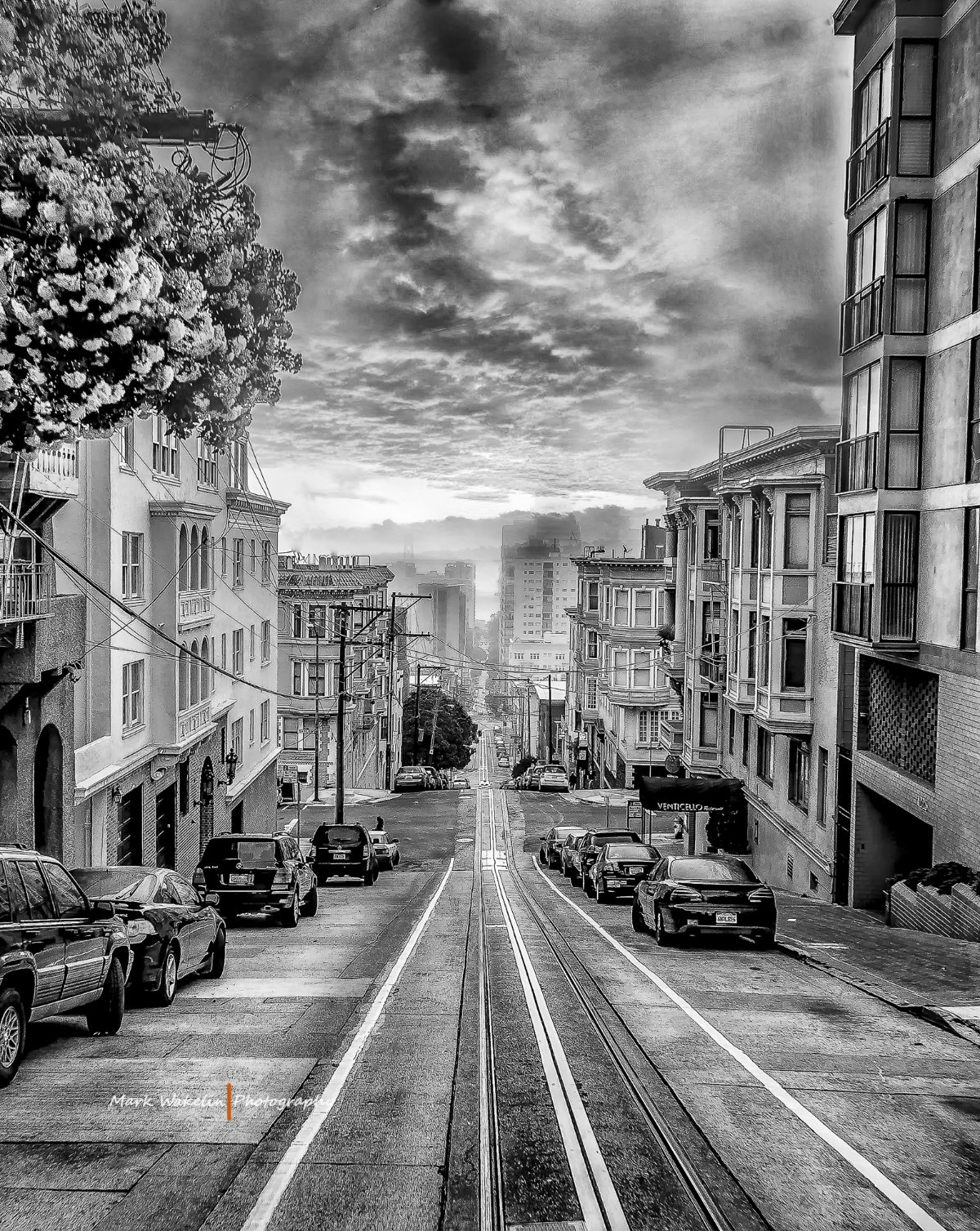 Black and white image of a steep San Francisco street lined with parked cars and classic buildings, featuring cable car tracks running down the centre.