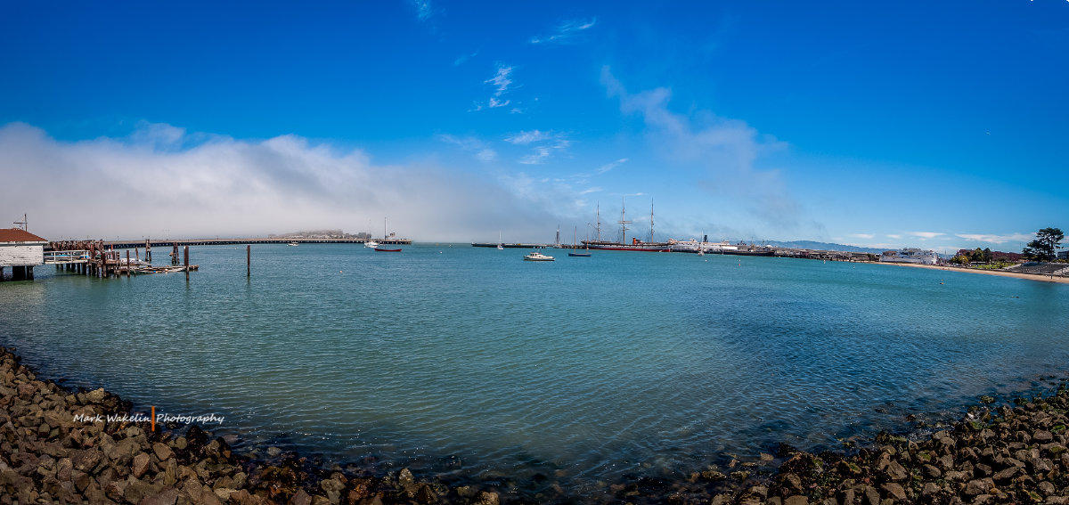 Panoramic view of a harbour with sailboats and a pier under a blue sky with scattered clouds.