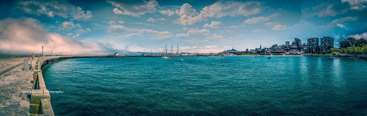 Panoramic view of San Francisco waterfront with a pier, sailboats on the water, and city skyline under a partly cloudy sky.