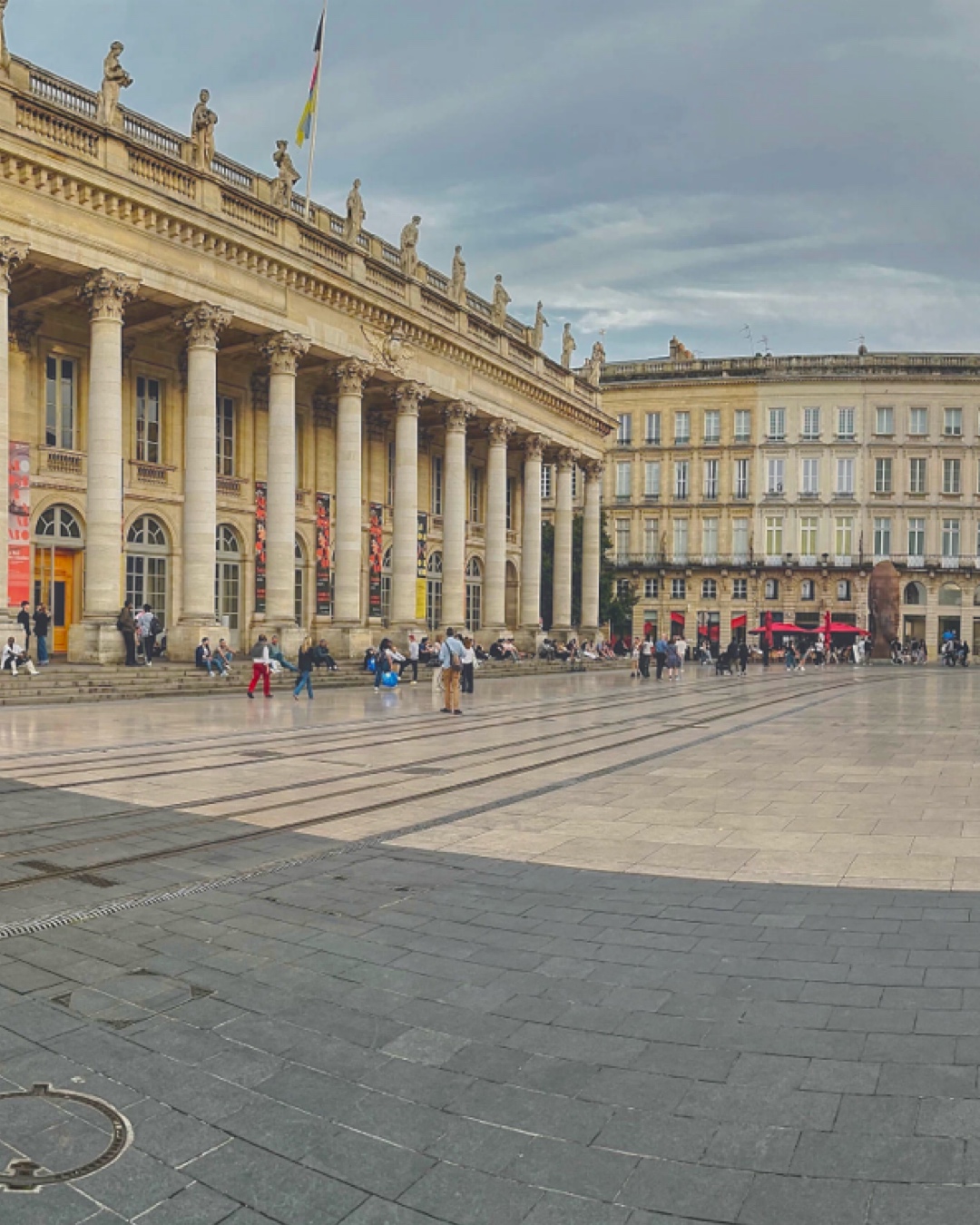 People gathered in the square in front of a large neoclassical building with tall columns and statues on the roof, under a cloudy sky.